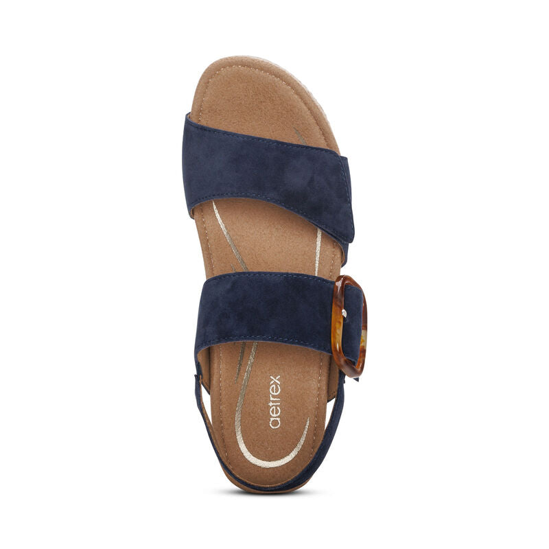 The Aetrex Women's Ashley Espadrille Wedge Navy by AETREX WORLDWide INC features navy leather, two wide straps, a tortoiseshell buckle, tan cushioned footbed with arch support, open toe design, and a visible brand logo on the insole.