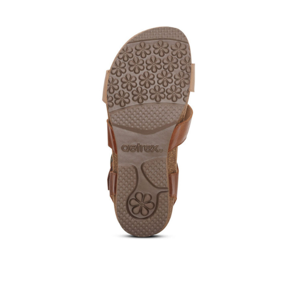 A Walnut Aetrex Women's Lilly Adjustable Quarter Strap Sandal by AETREX viewed from the bottom, showing its textured rubber sole with floral patterns, arch support, and AETREX branding embossed at the center.