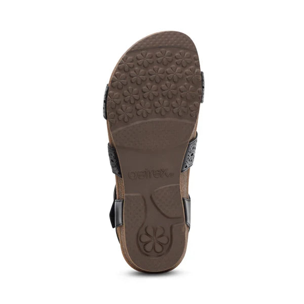 The image shows the bottom view of the Aetrex Women's Lilly Cutout Black sandal by AETREX WORLDWide INC, featuring a brown sole with a floral textured pattern and arch support detail. Black straps can be seen on the sides of the sandal.