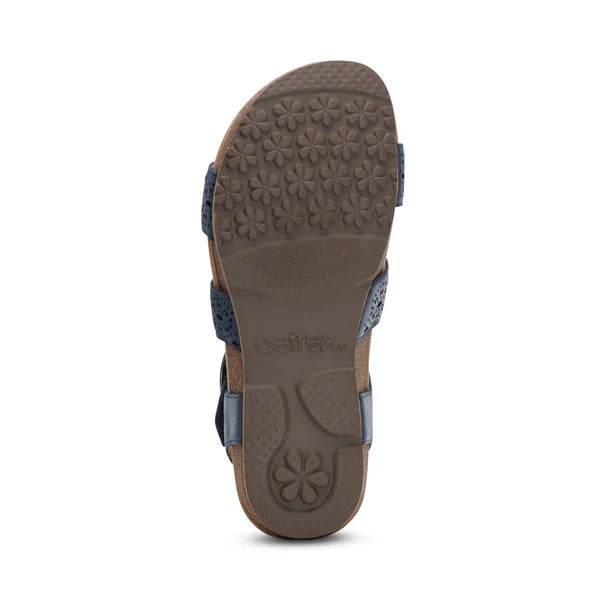 The image shows the bottom view of the Aetrex Women's Lilly Cutout Navy sandal by AETREX WORLDWide INC, featuring a flower-patterned textured rubber sole, blue straps, a contoured memory foam footbed, and arch support.