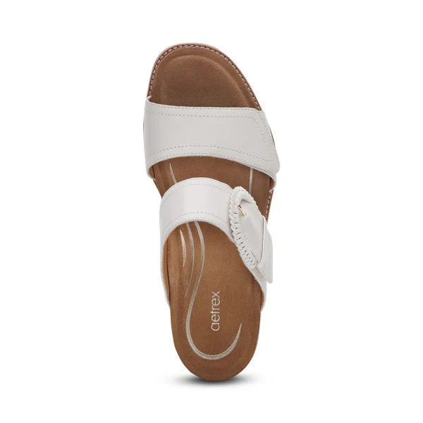 The Aetrex Women's Tatiana Ivory by AETREX WORLDWide INC is a white slip-on sandal with two wide straps and a cushioned tan footbed, ideal for plantar fasciitis. Subtle stitching accents the design, with Aetrex branding on the insole.