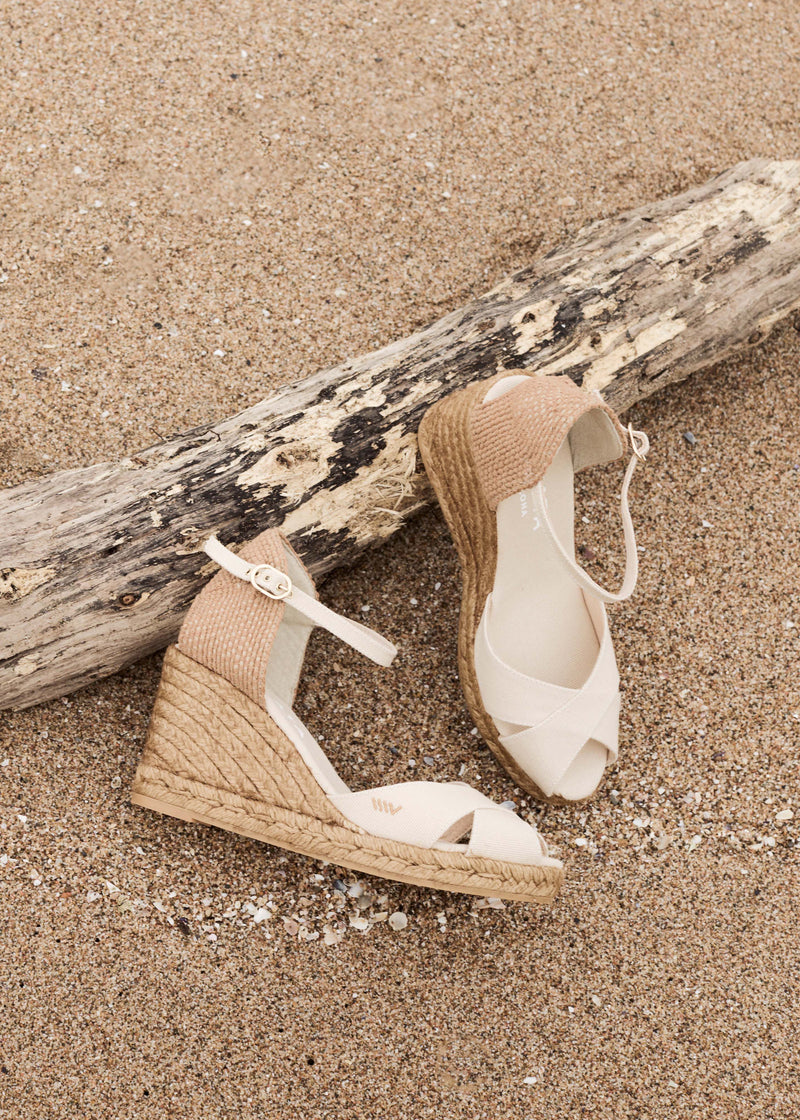 Side view of beige slingback espadrille wedges with crisscross open-toe straps and jute-wrapped wedge soles on beach sand next to driftwood.