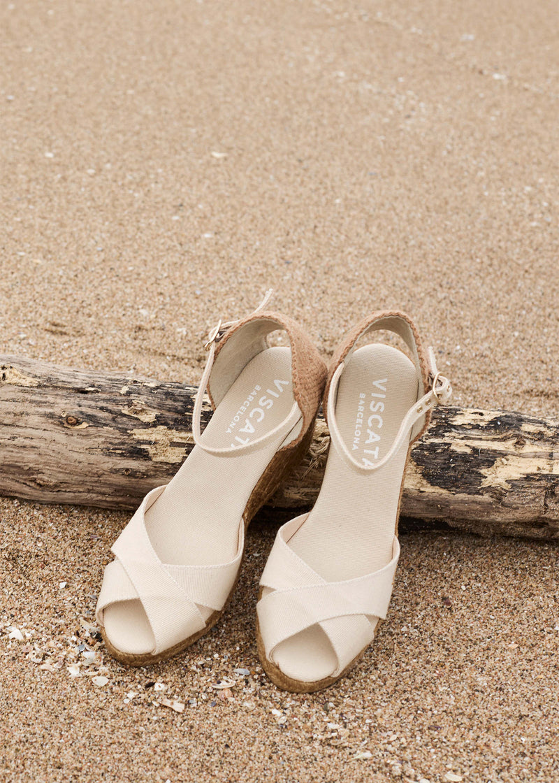 Two beige ankle-strap espadrille wedges with crisscross open-toe straps and jute-wrapped soles rest on beach sand beside a piece of driftwood.