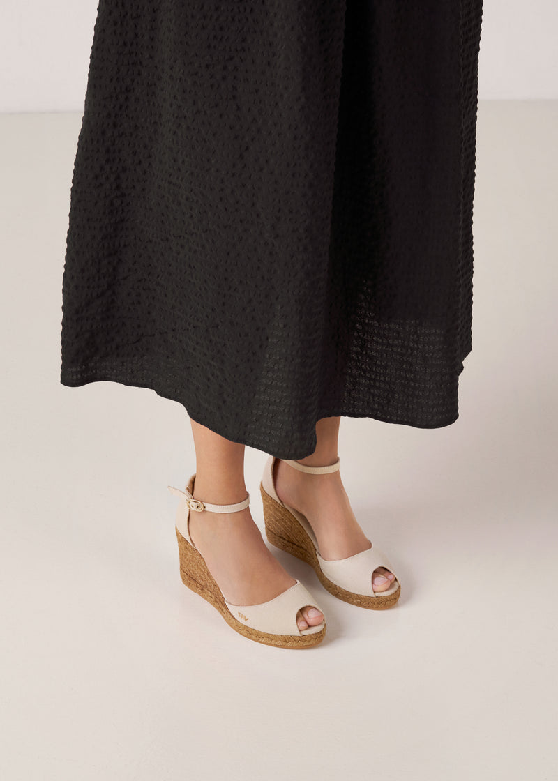 Beige canvas ankle-strap wedge espadrilles with a natural jute platform, worn with a black midi skirt