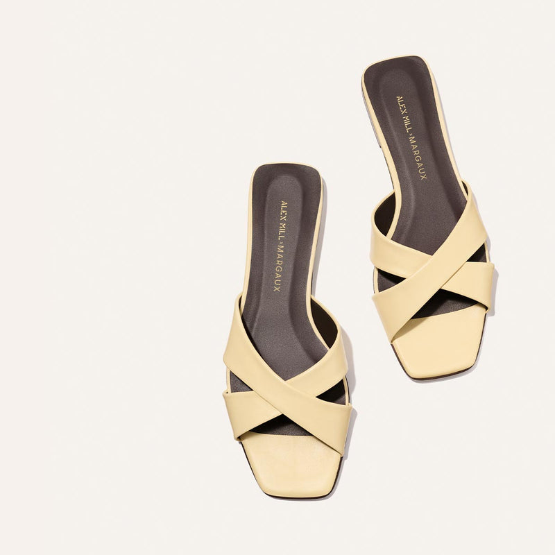 The Margaux Alex Mill x Margaux Selene Flat Sandal in Butter Nappa features pale yellow crisscross leather straps, square toes, and dark insoles with gold branding, all set against a plain background.