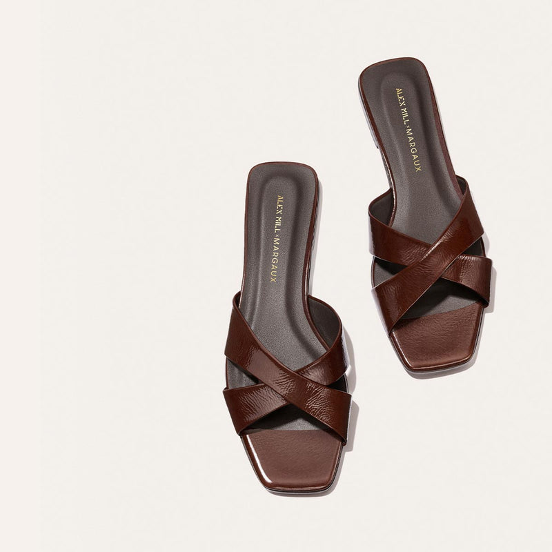 The Margaux Alex Mill x Margaux Selene Flat Sandal in Espresso Crinkle Patent features dark brown, crisscross leather straps, square toes, and a black insole with gold text—ideal for a chic summer look.
