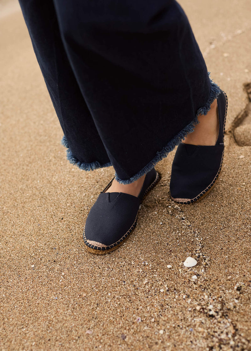 Close-up of a woman walking barefoot on sand in navy Barceloneta slip-on flats, paired with dark denim trousers. The simple lines and natural textures evoke an effortless coastal spirit.