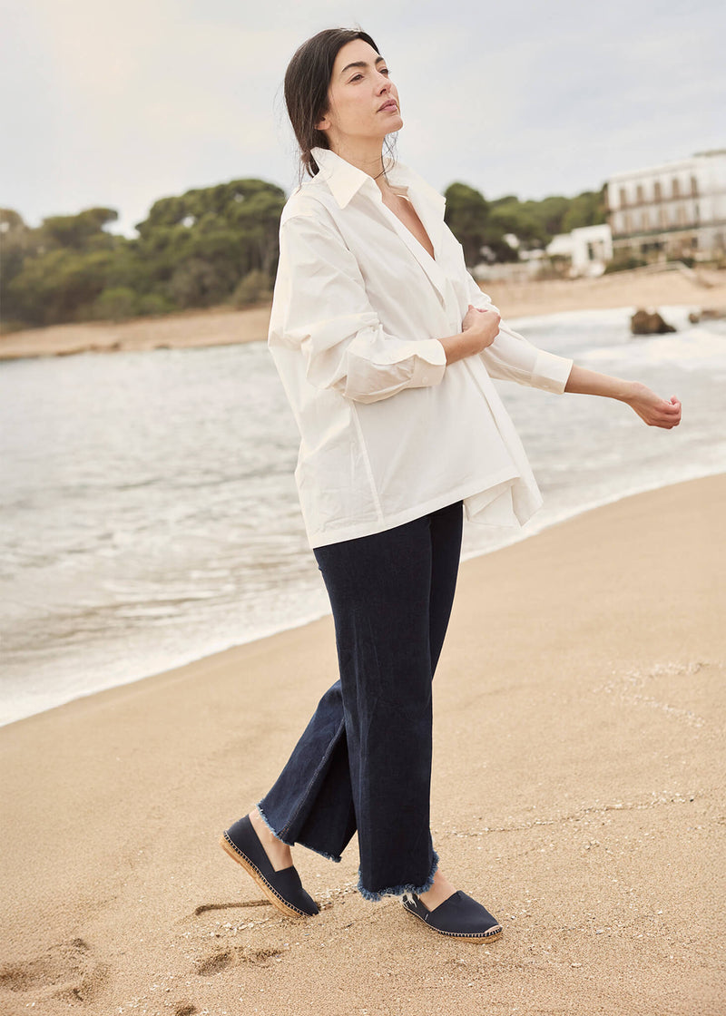 Woman walking along the beach wearing an oversized white shirt, dark denim trousers, and Barceloneta navy blue canvas slip-on espadrilles, capturing a relaxed coastal spirit.