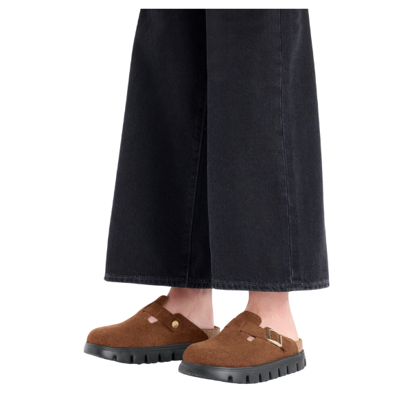 A person stands against a white background wearing wide-leg black pants and Birkenstock Women's Boston Chunky Dark Tea clogs by BIRKENSTOCK, featuring brown suede, soft footbed, black soles, and a gold buckle.