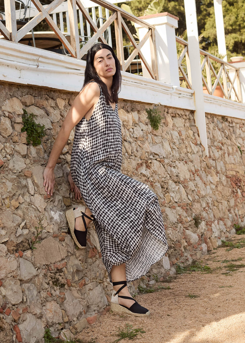 Woman sitting gracefully along a stone wall under the Mediterranean sun, wearing Escala black lace-up wedge espadrilles and a flowing gingham dress. A serene moment of timeless summer style.