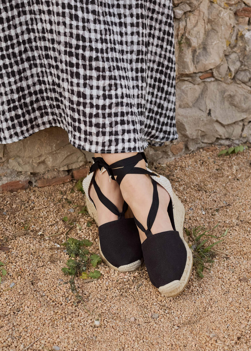 Close-up of a woman wearing Escala black lace-up wedge espadrilles, paired with a flowing black-and-white gingham dress. The natural stone backdrop and earthy ground highlight a refined yet relaxed Mediterranean spirit.
