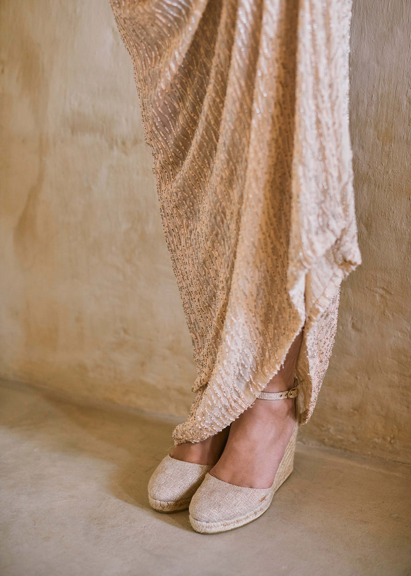 Champagne gold espadrille wedges with ankle straps styled beneath a flowing textured evening dress in a warm Mediterranean interior.