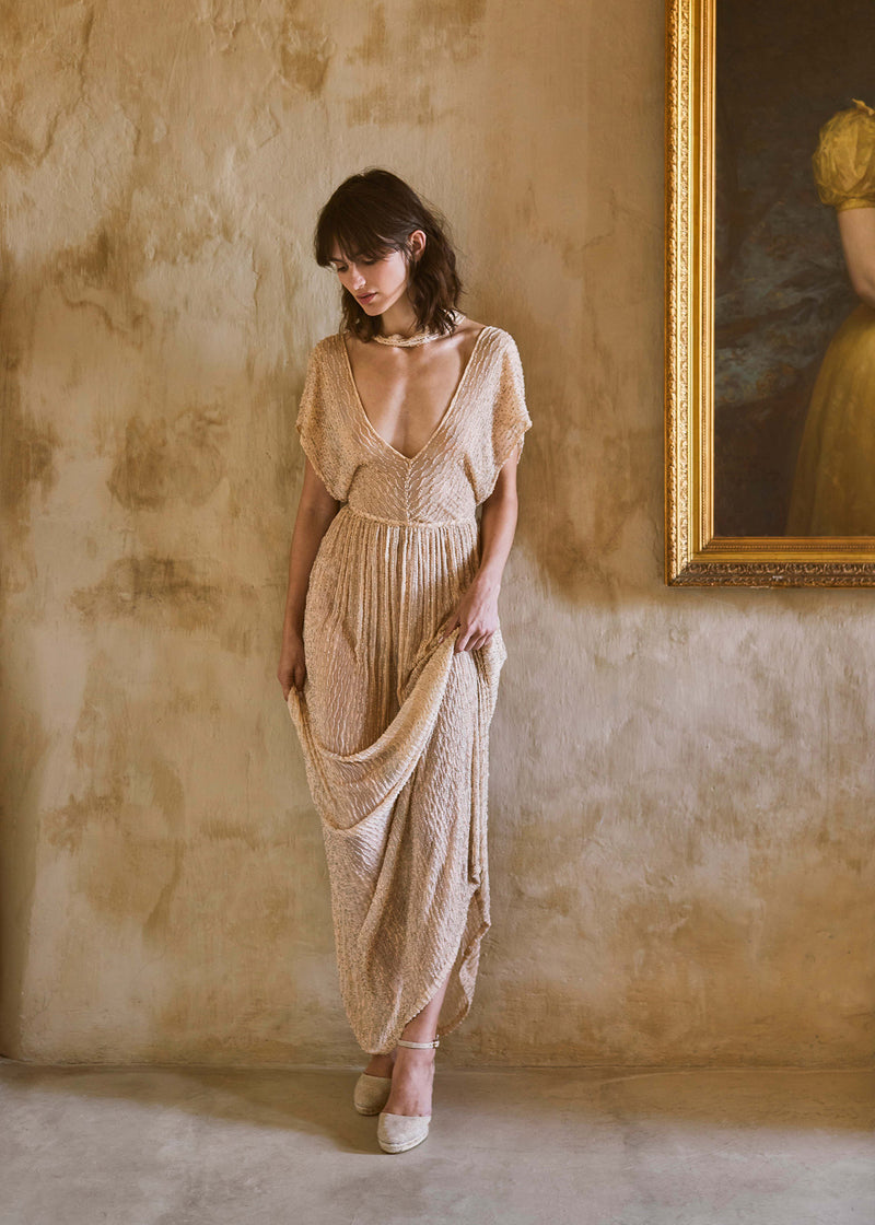 Woman wearing champagne gold espadrille wedges styled with a flowing textured dress in an elegant Mediterranean interior.