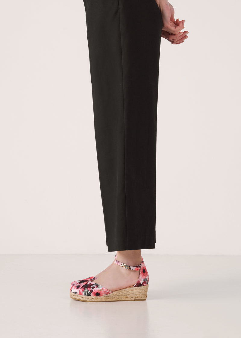 Side view of floral print wedge espadrilles with ankle strap, open toe, and a natural braided jute wedge, styled under black cropped trousers in a clean studio setting