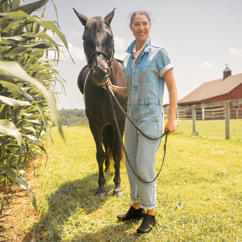 A farmgirl in denim coveralls wearing black boots while working with horses.