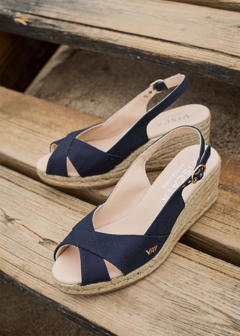 Two navy slingback espadrille wedges with crisscross open-toe straps and jute-wrapped wedge soles rest on weathered wooden steps.