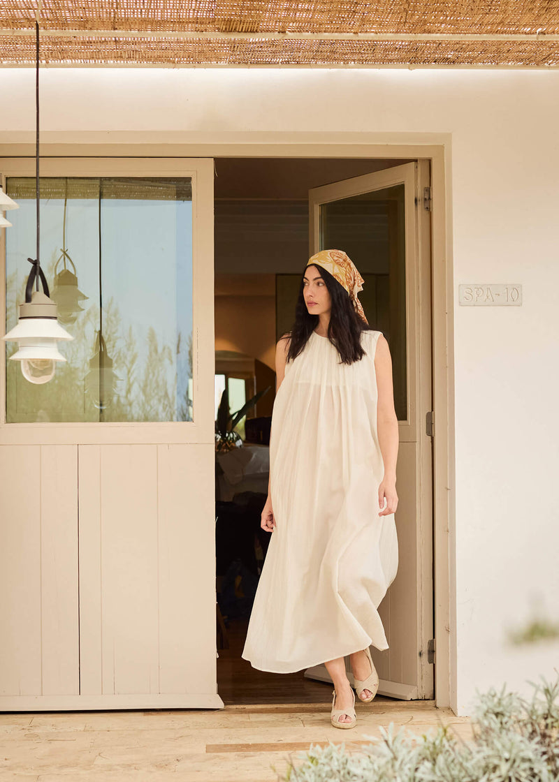 Woman stepping outside into the sunlight wearing a flowing cream dress and Llansa gold canvas slingback wedge espadrilles, paired with a printed headscarf.
