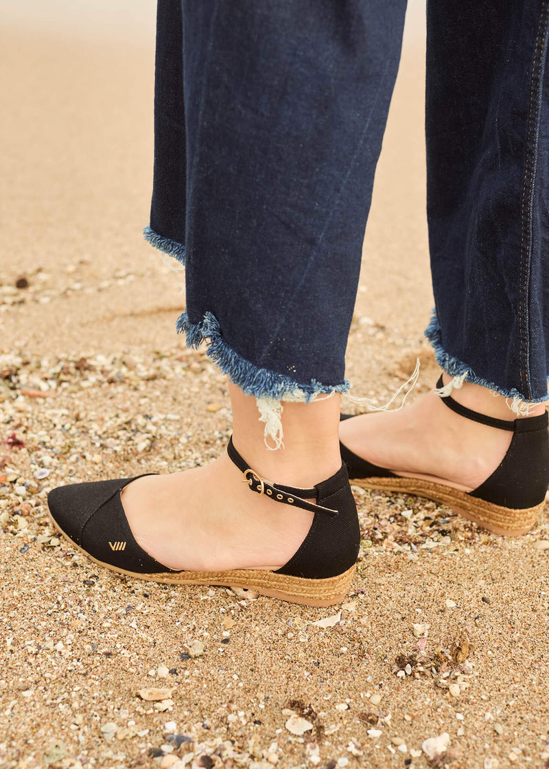 Close-up of a woman standing on the shoreline, wearing Montroig black ankle-strap flat espadrilles with frayed-hem denim trousers. The soft sand and casual silhouette capture an effortless coastal charm.