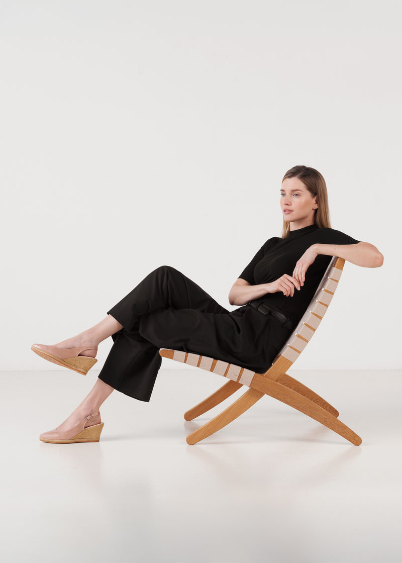 Woman seated in a woven lounge chair wearing blush pink leather wedge espadrille sandals, styled with a black outfit in a clean studio setting