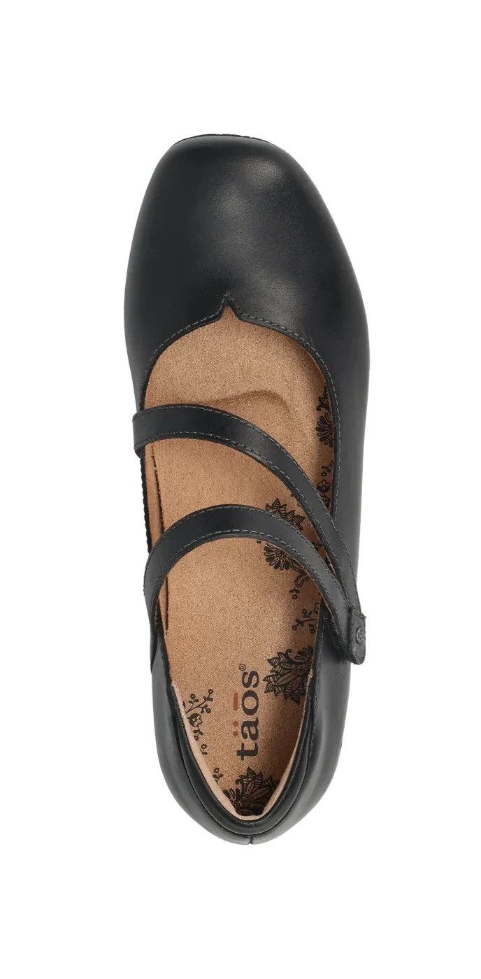 The Taos Womens Banter Black by TAOS is a luxe black leather Mary Jane flat with two straps, a tan insole featuring floral patterns and taos branding, plus arch & metatarsal support, shown from above on a white background.