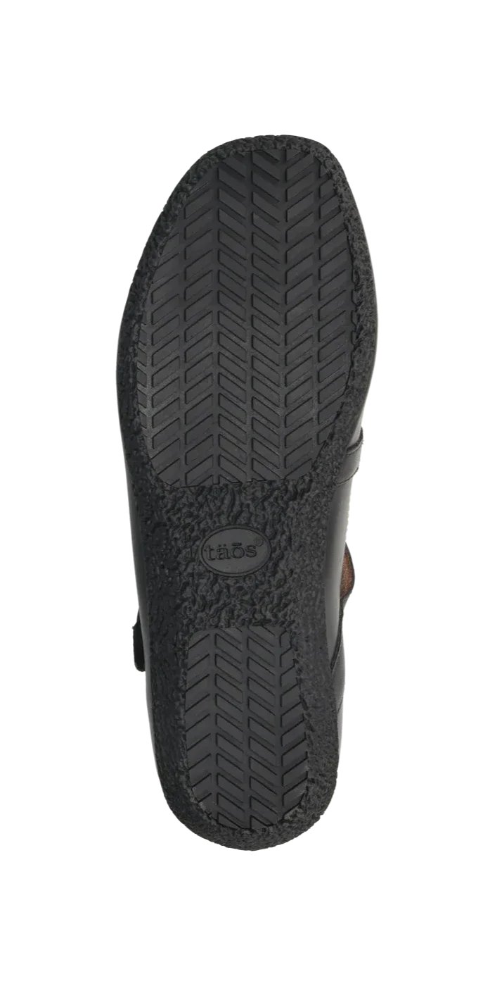 The image displays the black rubber sole of the TAOS Womens Banter Black flat, featuring a herringbone tread and center Taos logo, emphasizing its arch and metatarsal support for all-day comfort.