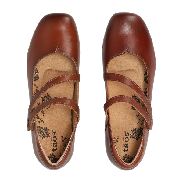The Taos Women's Banter Cognac are brown leather Mary Jane shoes with two diagonal straps, floral-patterned tan insoles, arch support for comfort, and the TAOS brand name printed inside.