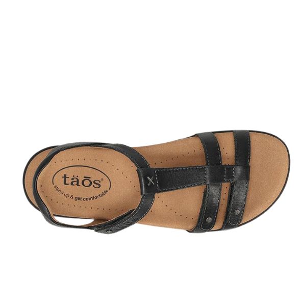 The Taos Womens Trophy 2 Black Emboss sandal by TAOS features a tan cushioned footbed with arch support, the taös logo, and “step up & get comfortable” inside—combining comfort and style for all-day wear.