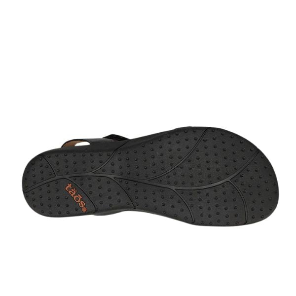 The image shows the black textured rubber sole of a TAOS Womens Trophy 2 Black Emboss sandal, featuring small circular bumps, curved grooves for grip, and the TAOS brand name in orange near the heel.