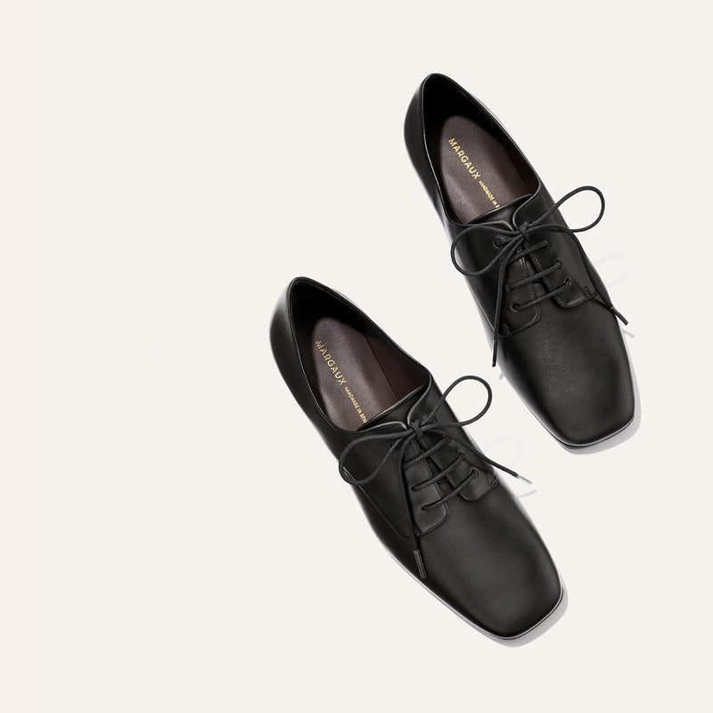 A pair of Margaux's The Billie Derby in black nappa leather, featuring square toes and lace-up design, displayed on a light-colored background.