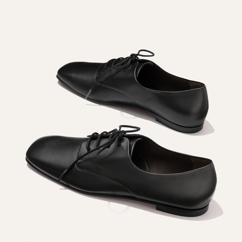 A pair of Margaux’s The Billie Derby shoes in Black Nappa leather is displayed on a light background, with one shoe positioned slightly above the other.