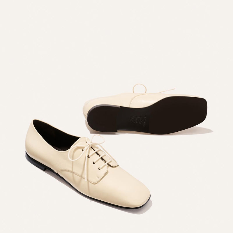 A pair of Margaux's The Billie Derby - Vanilla Nappa shoes: cream-colored lace-up leather with black soles, one shoe lying on its side to display the sole, the other standing upright.