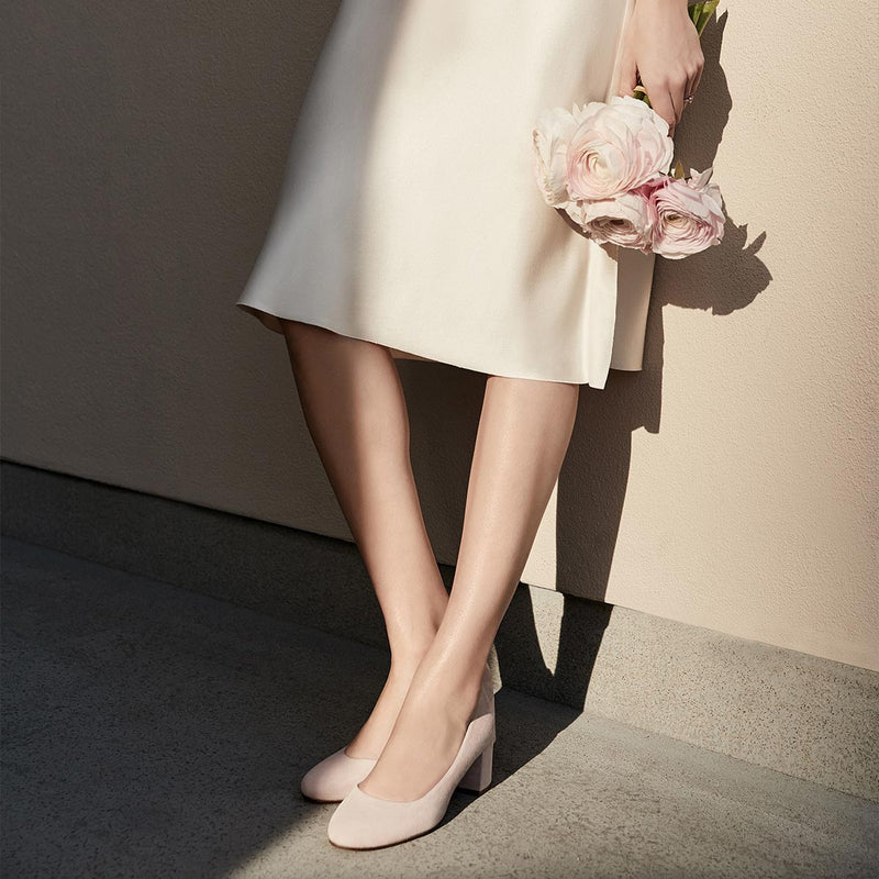 A woman in a light dress stands against a beige wall, holding pale pink flowers. Only her legs and dress show, paired with bespoke bridal shoes. Her look is completed with The Bridal Atelier E-Gift Card from Margaux.