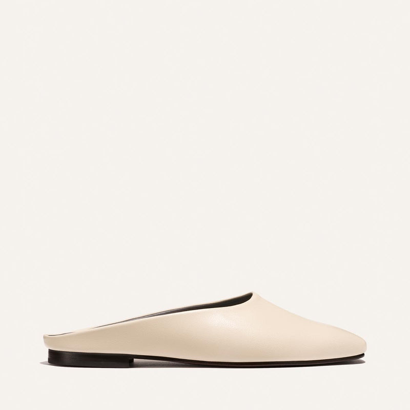 The Clara - Bone Nappa by Margaux is a minimalist, cream-colored slip-on mule with a closed toe and flat black sole, shown from the side against a plain white background.