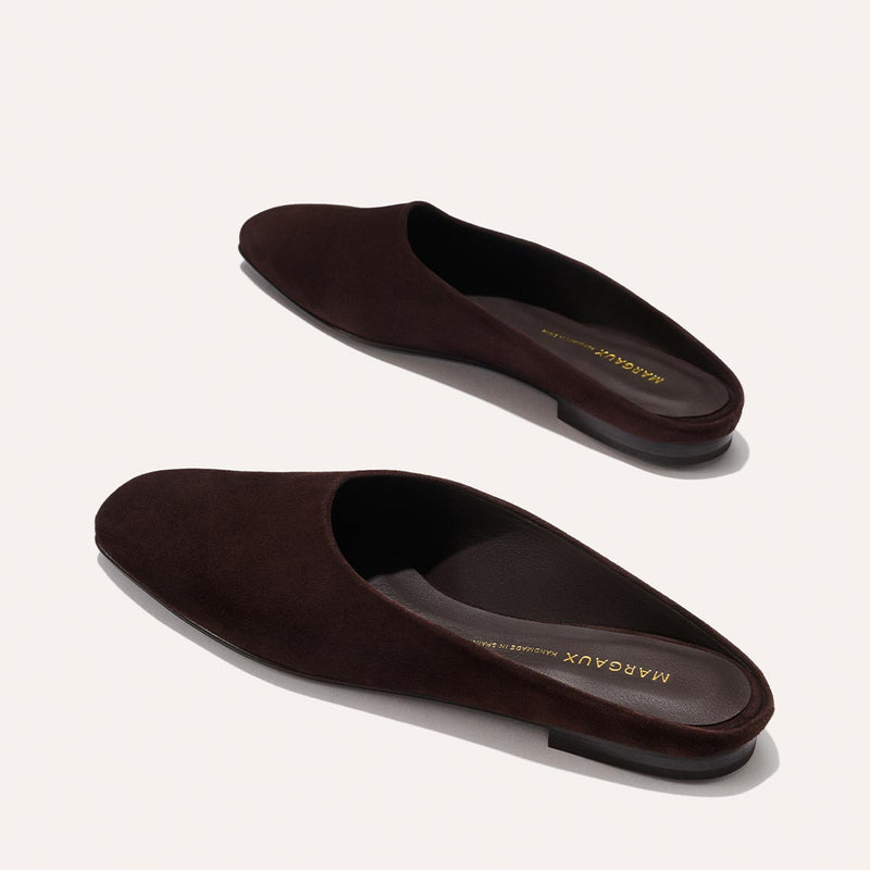 The Clara - Espresso Suede by Margaux are dark brown suede slip-on mules with flat soles and a minimalist, slipper-inspired design. Shown on a light background, the brand name Margaux is visible inside the shoes.