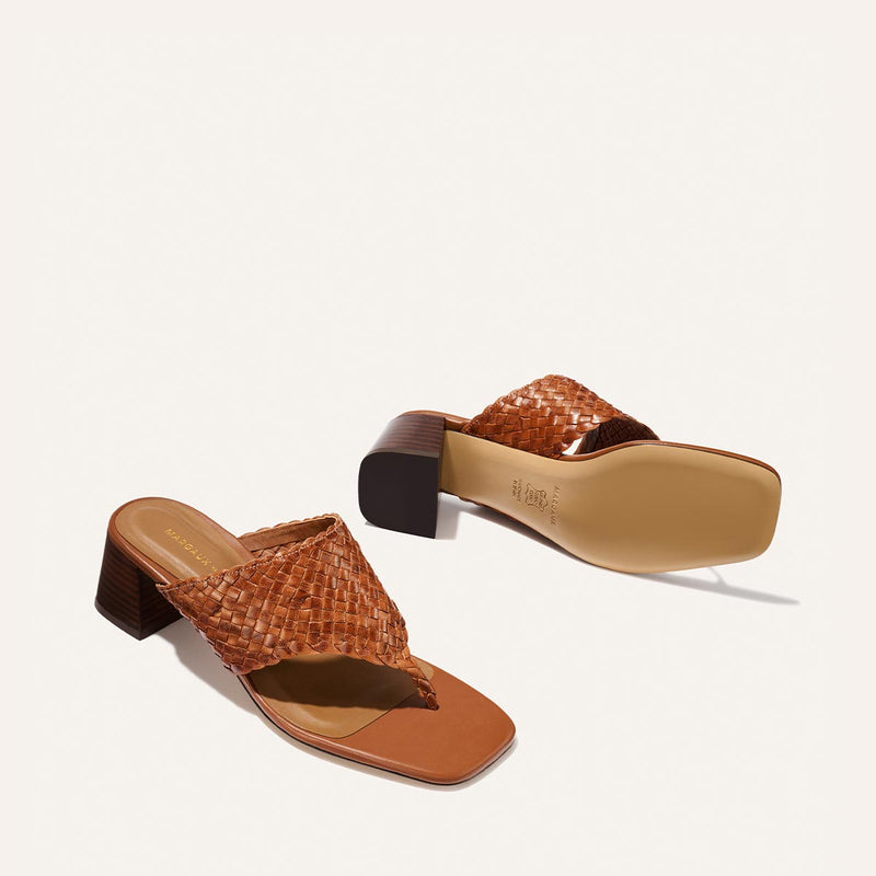 The Deia 50 - Saddle Woven Leather sandals by Margaux, featuring brown woven leather, block heels, a square toe, and thong straps, are shown on a light background—one upright and one on its side to display the sole.