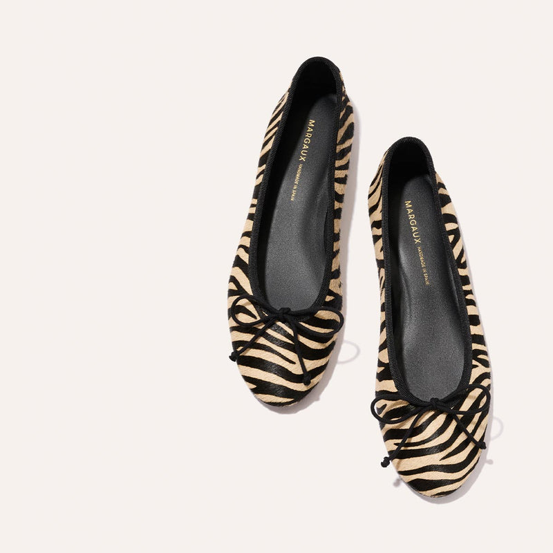 The Demi - Tan Zebra Haircalf ballet flats by Margaux, featuring comfortable zebra-print and black bow accents on the toes, are displayed against a light background.