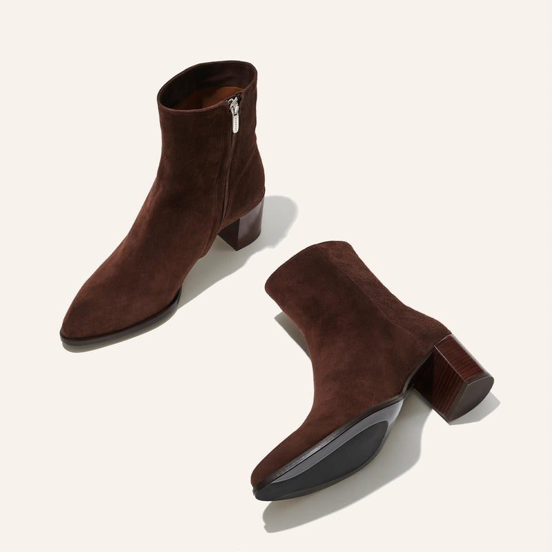 The Margaux Downtown Boot First Edition in Chocolate Suede is shown on a light background; one boot stands upright while the other lies on its side, highlighting the block heel, side zipper, and the sole.