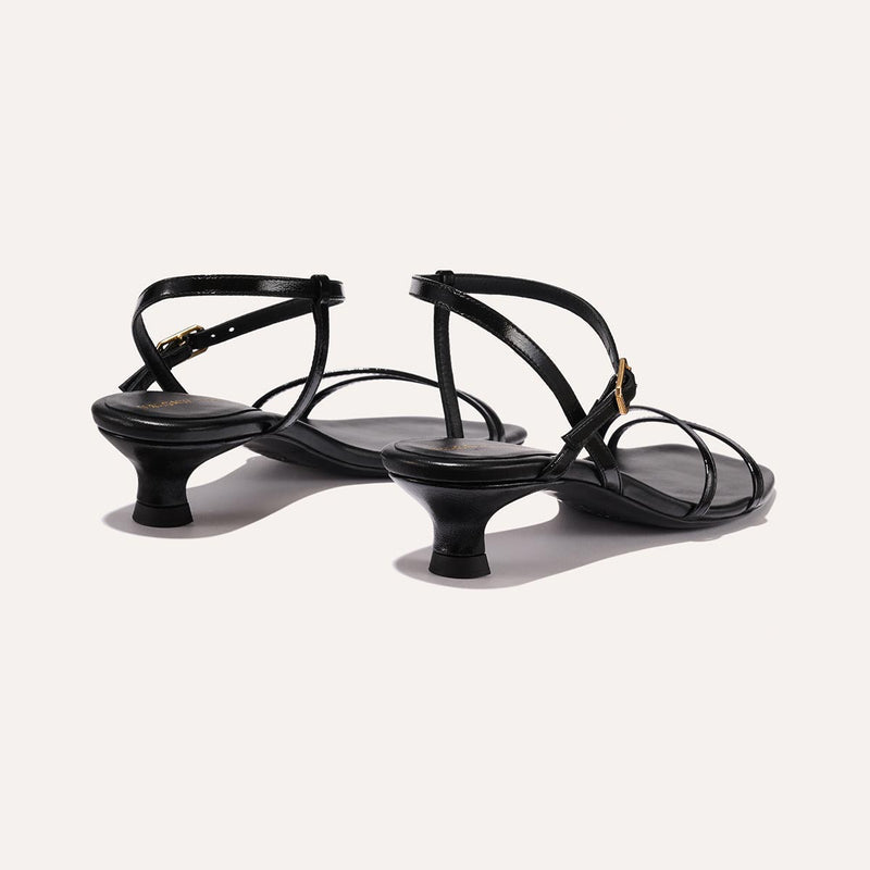 The Dylan Sandal in shiny Black Crinkle-Gloss Nappa, featuring asymmetrical straps around the ankle and toes and a walkable 1-inch stiletto heel.