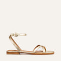 The Flat Sandal - Champagne by Margaux: a gold barely-there style with thin straps, an ankle strap with round buckle, flat brown sole, and foam-cushioned insole for comfort. Shown on a plain white background.