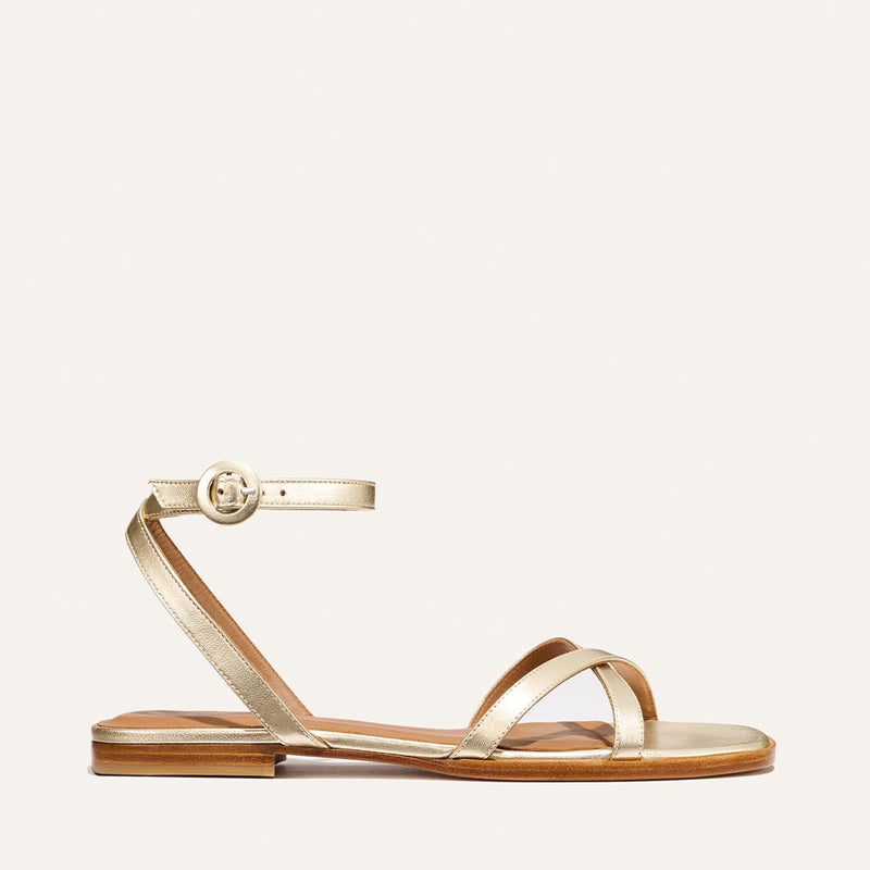 The Flat Sandal - Champagne by Margaux: a gold barely-there style with thin straps, an ankle strap with round buckle, flat brown sole, and foam-cushioned insole for comfort. Shown on a plain white background.