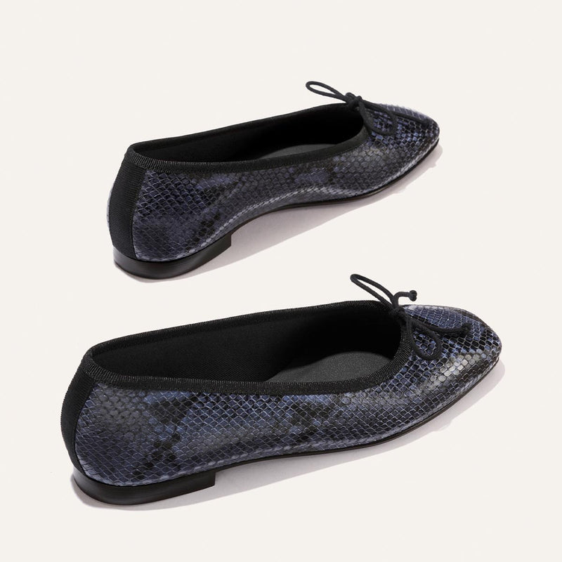 Margaux's The Fonteyn - Fumo Python Embossed flats feature a pointed toe, dark blue and black snakeskin pattern, black trim, and small black bows on the front, shown on a light background.