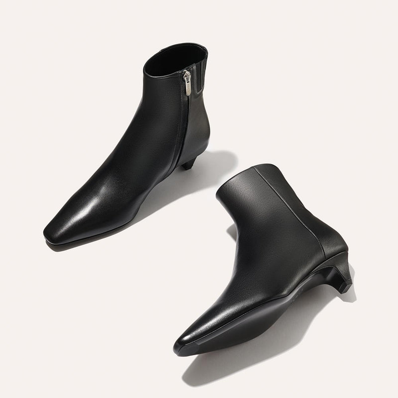 The Inga - Black Calf by margaux-development: black ankle boots with pointed toes, short heels, and side zippers—NYC-inspired essentials. Displayed with one boot upright and the other on its side against a light background.