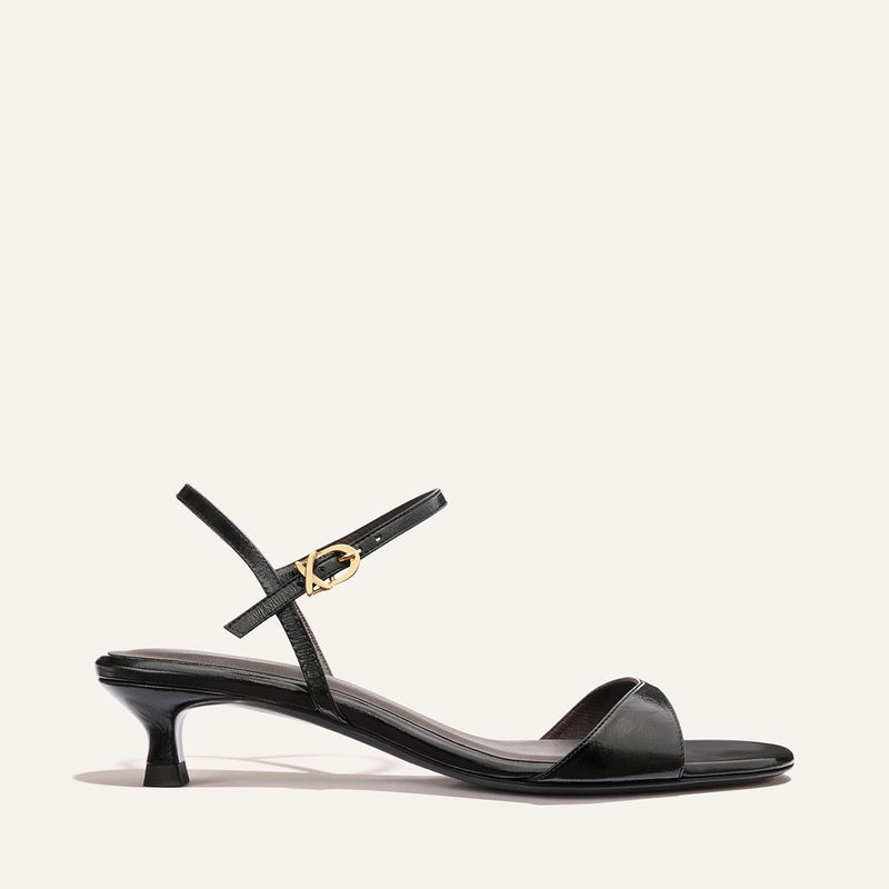 The Lotto 35 - Black Crinkle-Gloss Nappa by Margaux is a black strappy sandal with a low kitten heel, thin ankle strap with a gold buckle, and minimalist design shown against a plain light background.