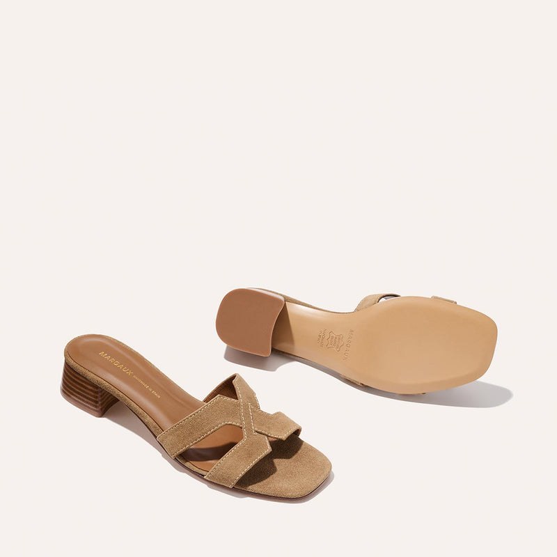 The MX 35 - Biscotti Suede by Margaux: tan open-toe heeled sandals with crisscross suede straps and a low block heel. Displayed on a light background, one sandal is upright while the other shows the sole.