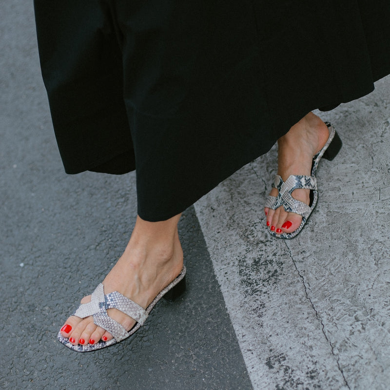 A person wearing a long black skirt and open-toed margaux-development The MX 35 - Natural Python Embossed sandals with block heels and red nail polish stands elevated on the edge of a street.