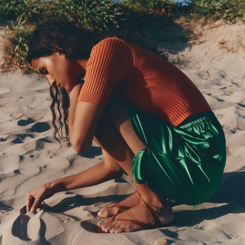 A woman with long wavy hair crouches on sandy ground, drawing in the sand amid summer greenery. She wears a short-sleeved orange top, green skirt, and Margaux’s The Palermo Sandal - Saddle Nappa with a cushioned insole.
