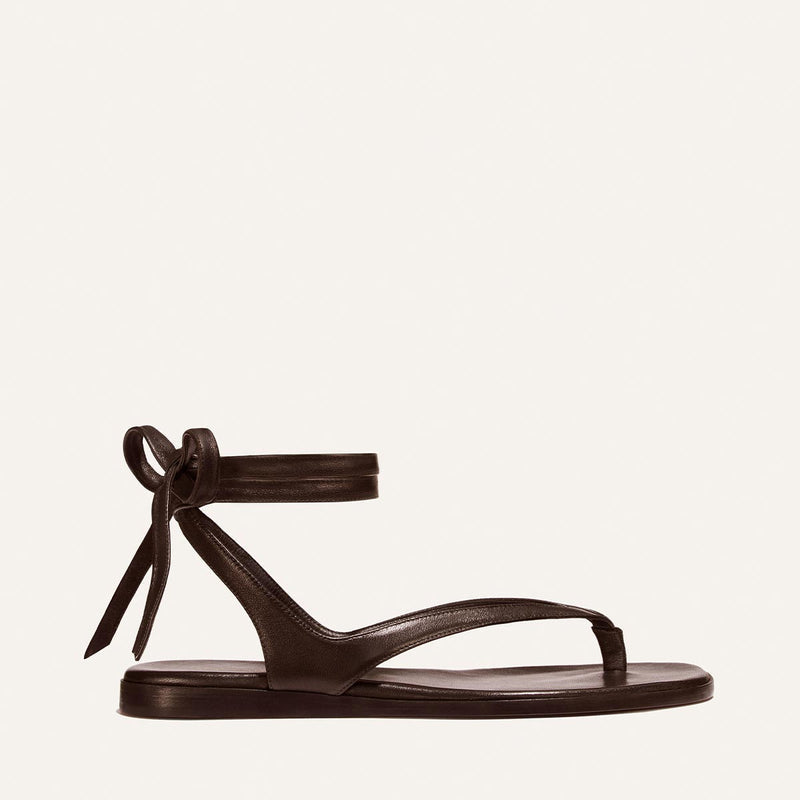 The Palermo Wrap Sandal - Espresso Nappa by Margaux features minimalist black leather, a flat sole, cushioned insole, toe loop, and wrap-around ankle straps tied in a bow—perfect summer style shown on a light background.