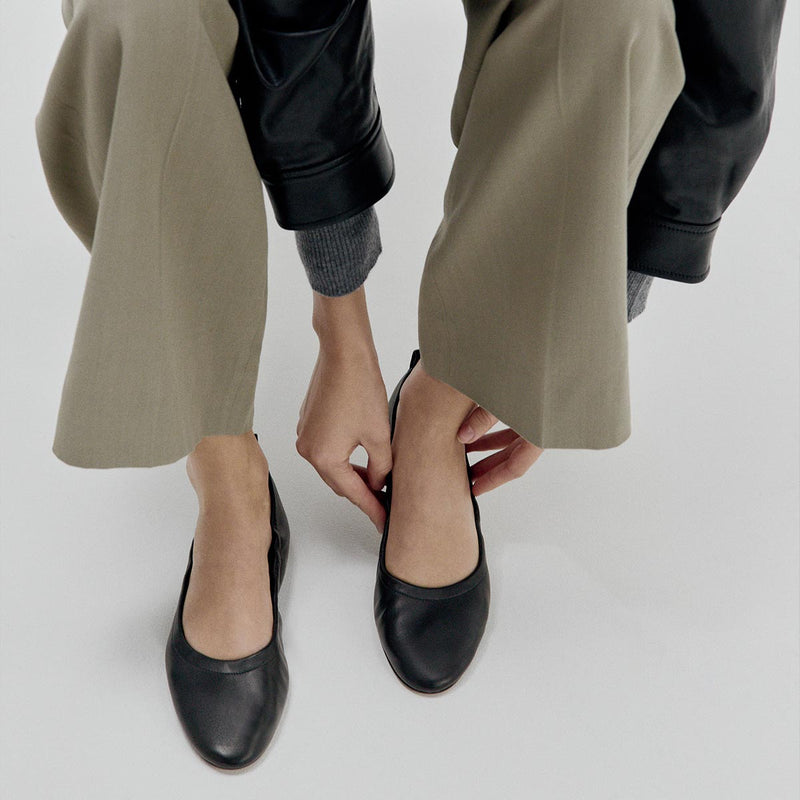 A person wearing taupe wide-leg pants and a black jacket adjusts Margaux's The Pascale - Black Nappa ballet flats on their feet, photographed from above against a white background.