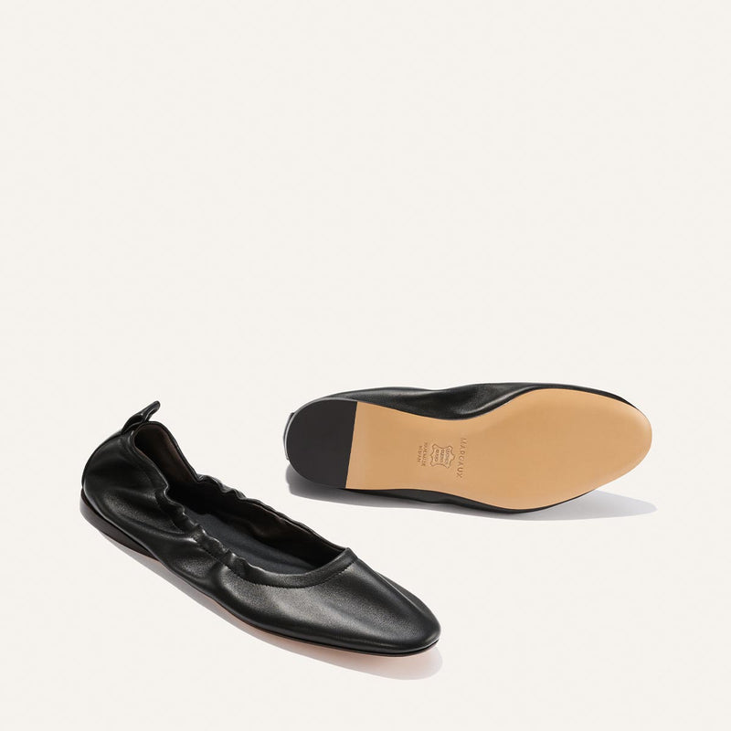 A pair of Margaux's The Pascale - Black Nappa ballet flats; one shoe shows the top view, while the other displays the light tan sole.