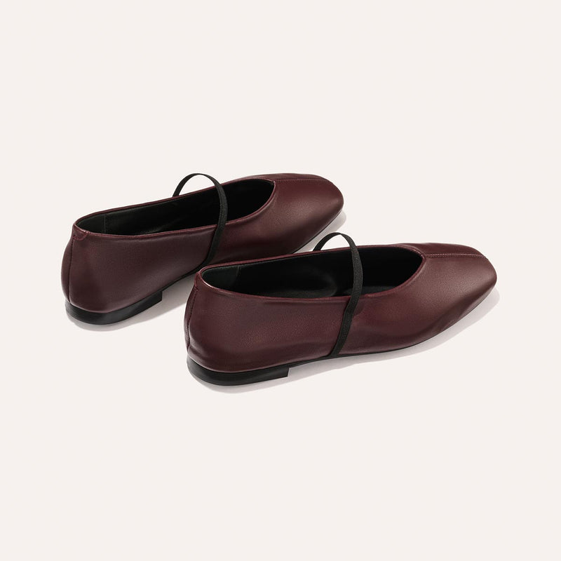 The Phoebe - Burgundy Nappa by margaux-development: burgundy ballet flats with black elastic straps, sacchetto construction for flexible comfort, rounded toe, and flat heel, shown on a plain light background.